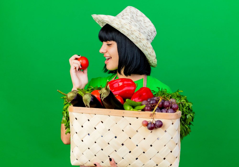 joyful-young-female-gardener-uniform-wearing-gardening-hat-holds-vegetable-basket-looks-tomato